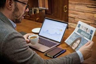 Man reading newspaper next to laptop and phone